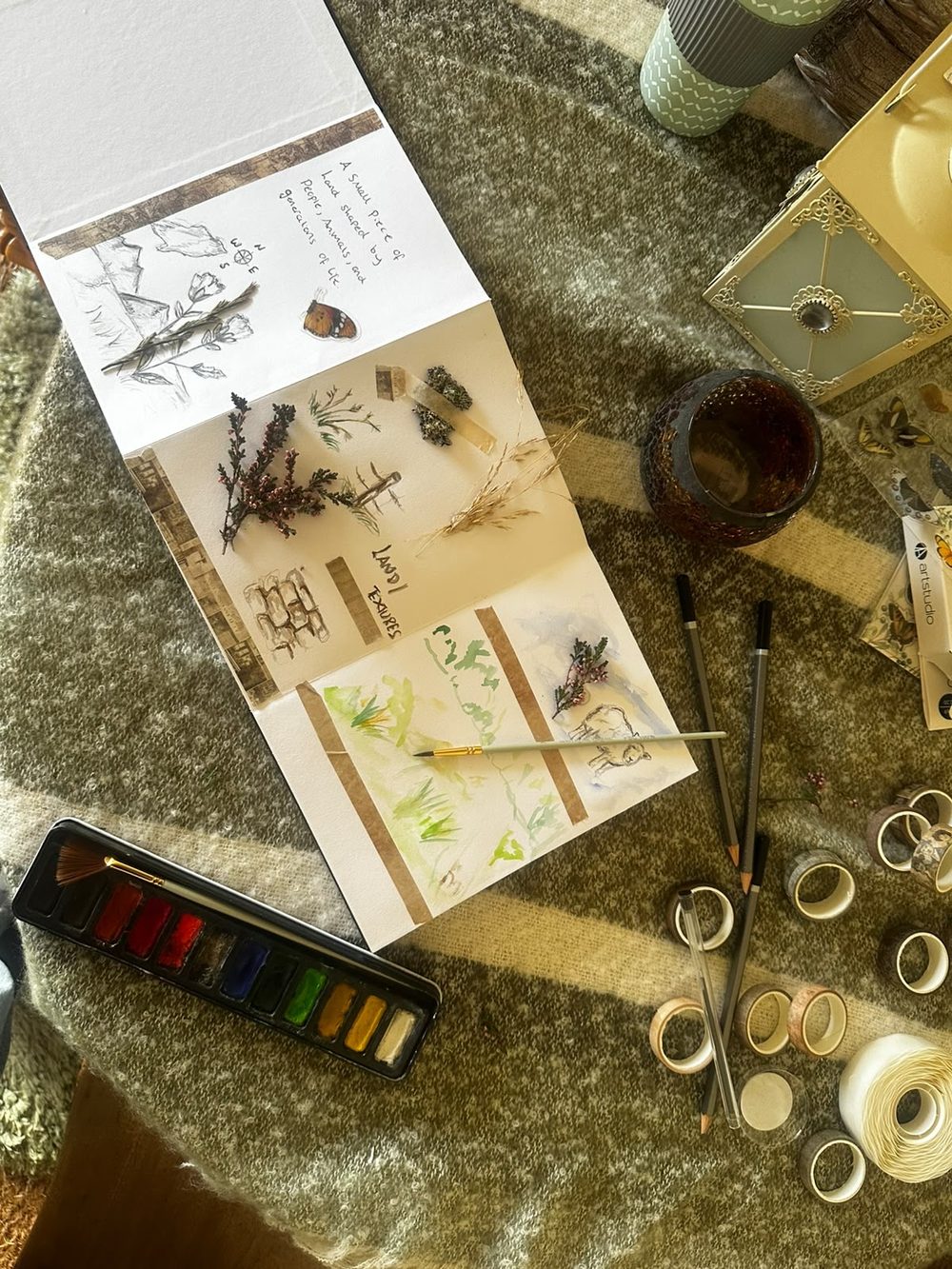 An open artist's book on a striped wool blanket, surrounded by watercolour paints, pencils and patterned tape