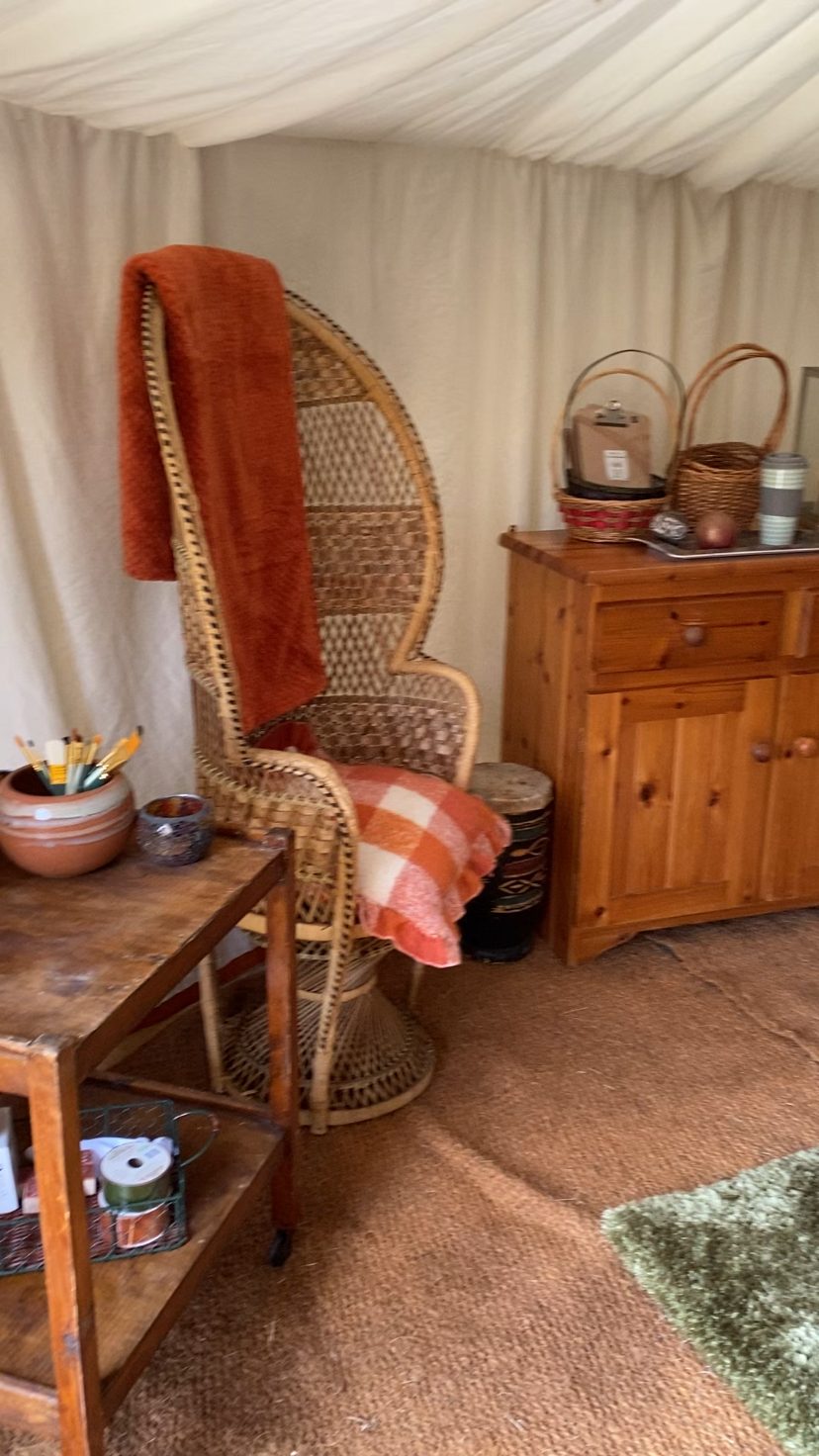 A wicker armchair draped with a fleece throw beside a trolley of craft supplies in the yurt